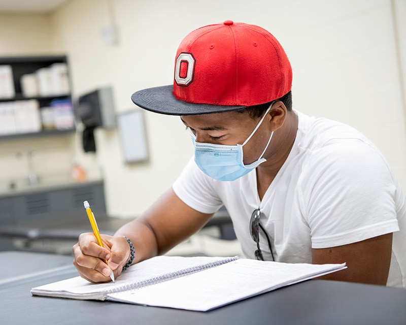 Student wear Ohio State hat writes at a desk